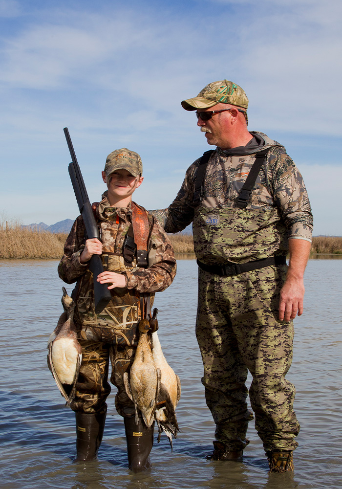 A boy and a man in camo clothes standing in a wetland. the boy, smiling, with a rifle and 3 ducks strapped to his belt.