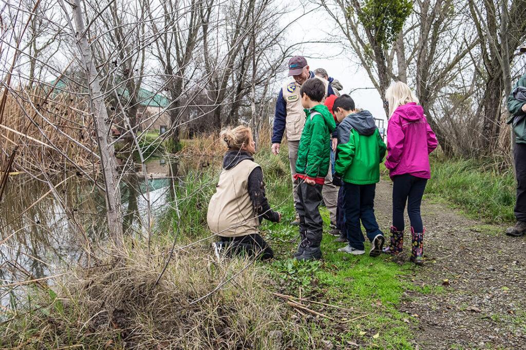 Children surrounding a docent on their knees on a small, wooded trail at the edge of a wetland area.